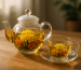 An elegant glass tea set with blooming flowers inside sits on a wooden table in natural light.