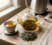 Loose leaf tea brewing setup with glass teapot, amber tea in ceramic cup, and scattered dry tea leaves on wood